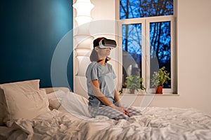 Young girl sits on bed in virtual reality glasses. Woman watching 3d movie in special helmet