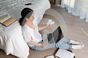 A young girl sits on the floor near the bed with a laptop and holds a paper airplane in her hands. Top view. The concept