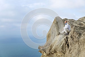 A young girl sits on a high cliff by the sea on a windy day. Cute blonde in a white jacket. Active recreation and travel