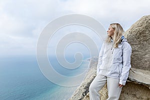 A young girl sits on a mountain top high above the sea on a sunny day. Cute blonde in a white jacket and sweatpants. Active