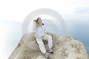 A young girl sits on a mountain top high above the sea on a sunny day. Cute blonde in a white jacket and sweatpants. Active