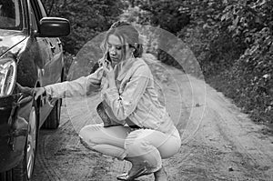 A young girl sits near a car with a broken wheel on the road and calls the rescue service to have her car repaired