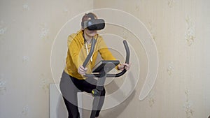 A young girl sits and pedals on an exercise bike at home in virtual reality glasses. A girl in yellow sportswear. Home