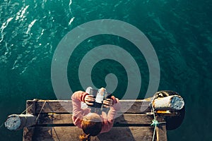 Young Girl Sits On The Pier And Looks At Sea Through Binoculars, Top View. Adventure Vacation Discovery Travel Concept