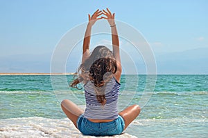 Young girl sits on the shore of the Dead Sea in Israel, view from behind. the brunette stretches her arms up