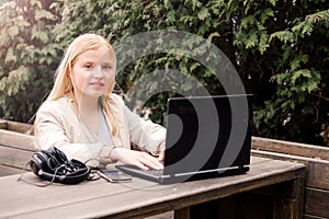 A young girl sits on the street in a cafe with a laptop, headphones and a phone lie next to the table