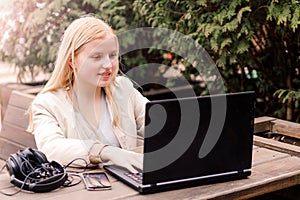 A young girl sits on the street in a cafe with a laptop, headphones and a phone lie next to the table