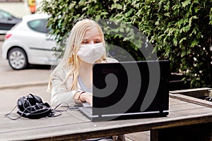 A young girl sits on a street in a cafe with a protective mask of white color. Girl looking at a laptop