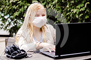 A young girl sits on a street in a cafe with a protective mask of white color. Girl looking at a laptop