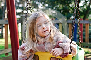 A young girl sits on a swing in a retro-styled playground, her expression is thoughtful and introspective.
