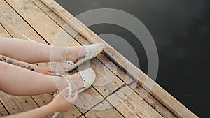 A young girl sits on a wooden bridge near the river and takes off her shoes. Close-up