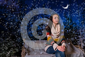 Young girl sitting on a bench or a swing in the evening in a snow-covered park with spruce trees, wearing warm woolen sweater