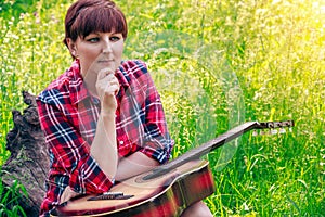 Young girl sitting on the grass in the field and plays the guitar. Beautiful nature at bright sunny summer day.