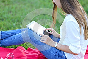 Young girl sitting on the grass in the park and works at a laptop and eating fast food
