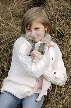 Young girl sitting on hay, smiling and holding a pig in his smiles. Lifestyle portrait