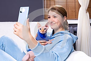 A young girl sitting at home on the couch makes a selfie with a bald cat. Sphinx with blue eyes looks into the camera of the phone