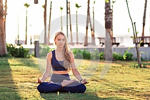 Young girl sitting in Lotus pose practicing Yoga in the park near seafront line at sunset sunrise