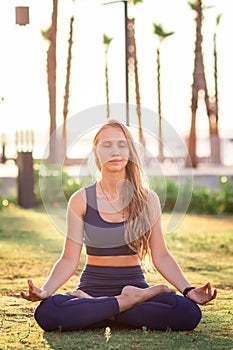 Young girl sitting in Lotus pose practicing Yoga in the park near seafront line at sunset sunrise