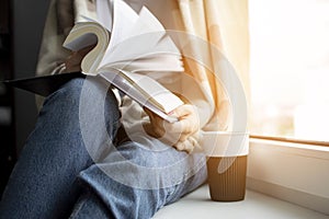 Young girl is sitting near the window, covered with a blanket, she is reading a book and drinking tea, close-up