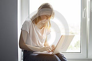 Young girl sitting near the window with a laptop, she uses the computer at home on the windowsill, a woman freelance, copy space