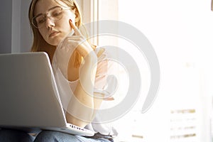 Young girl sitting near the window with a laptop, she uses the computer at home on the windowsill, a woman freelance, copy space