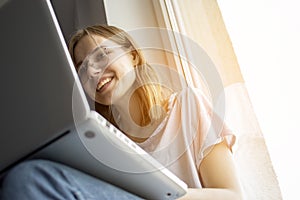 Young girl sitting near the window with a laptop, she uses the computer at home on the windowsill, a woman freelance, copy space