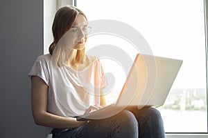 Young girl sitting near the window with a laptop, she uses the computer at home on the windowsill, a woman freelance, copy space