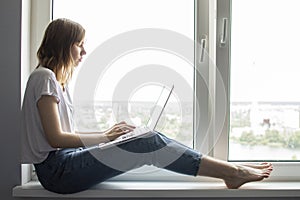 Young girl sitting near the window with a laptop, she uses the computer at home on the windowsill, a woman freelance, copy space