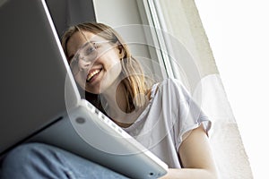 Young girl sitting near the window with a laptop, she uses the computer at home on the windowsill, a woman freelance, copy space