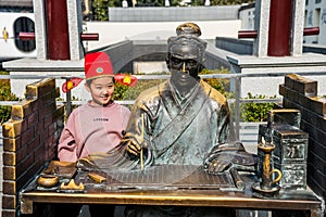 A young girl sitting next to the statue of Acient Chinese Scholar  writing on the bable  on the bamboo book