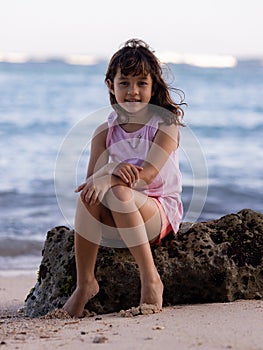 Young girl sitting on the rock near the ocean. Smiling girl. Happy childhood. Spending time on the beach. Vacation in Asia.