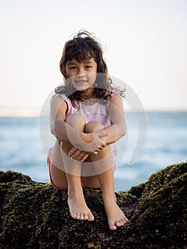 Young girl sitting on the rock near the ocean and smiling. Happy childhood. Spending time on the beach. Cute little girl. Vacation
