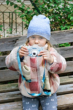 A young girl is sitting on a rustic wooden bench in a park, taking pictures with a blue camera.