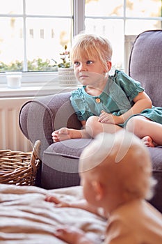 Young Girl Sitting On Sofa In Lounge At Home Watching TV As Baby Brother Plays In Foreground