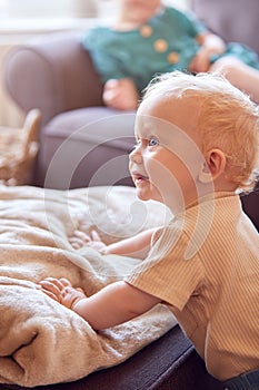 Young Girl Sitting On Sofa In Lounge At Home Watching TV As Baby Brother Plays In Foreground