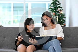 Young girl sitting on sofa and using digital tablet doing homework online during extra curricular classes with her mother. photo
