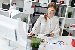 A young girl is sitting at a table in the office, holding a calculator in her hand and typing on the keyboard.