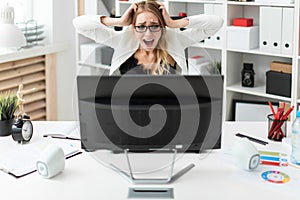 A young girl is sitting at a table in the office, holding hands behind her head and looking at the monitor.