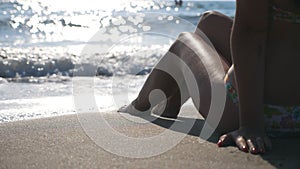 Young girl with slim legs sitting on tropical beach with splashing waves and enjoying vacation. Woman relaxing on coast