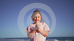 A young girl smiles playfully, clutching her hands together while standing on a sandy beach, with waves and clear blue