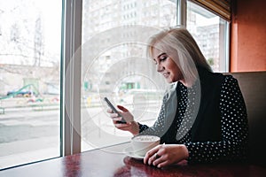 Young girl smiling blonde looks into the phone sitting in a cafe near the window with a cup of hot drink.