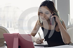 A young girl speaks on the phone, looking at the tablet screen and making notes in a notebook. The concept of freelancing, remote