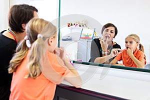 Young girl in speech therapy office. Mirror reflection of young girl exercising correct pronunciation with speech therapist.