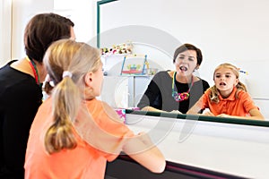 Young girl in speech therapy office. Mirror reflection of young girl exercising correct pronunciation with speech therapist.