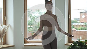 Young girl in sportswear does jump exercise in bright room with skipping rope