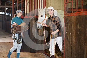 Young girl stable worker ready to saddle horse while Asian woman leading out of stall