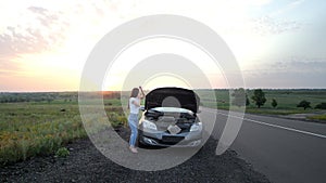A young girl is standing by a broken car on the highway.The girl looks under the hood of a broken car, trying to fix it