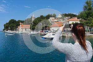 Young girl standing on the coast town taking a photo of adriatic sea with smart phone in Cavtat, Croatia. Red head pony tail