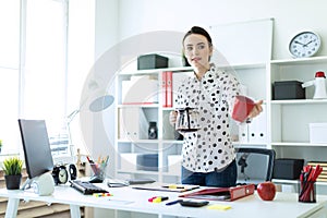 A young girl is standing in the office near the table, holding a kettle in her hand and holding out a cup.