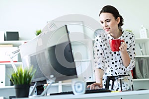 A young girl is standing in the office near the table, holding a red mug in her hand and typing on the keyboard.